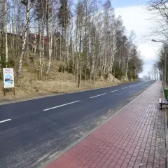 Droga w lesie, z tablicą "Uwaga! Sensacja!", sugerującą miejsce anomalii grawitacyjnej. Po prawej chodnik i ławka.