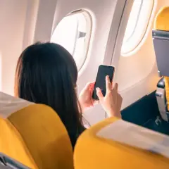 Kobieta w samolocie trzyma telefon, zastanawiając się, **co to tryb samolotowy**. Widok zza jej pleców na żółte siedzenia i okno.