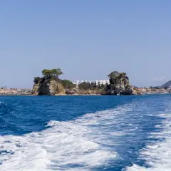 Idealna zakynthos pogoda sierpień: błękitne niebo, turkusowe morze i skaliste wysepki z drzewami. Widać łódź i plażę.