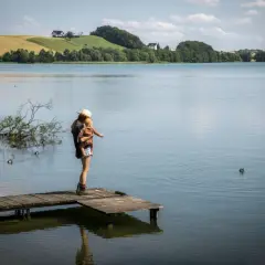 Kobieta z dzieckiem na rękach na pomoście, podziwiająca krajobraz jeziora i wzgórz. Szwajcaria Kaszubska atrakcje w pełnej krasie.