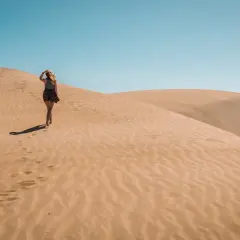 Kobieta spaceruje po piaszczystych wydmach Dunas de Maspalomas, zostawiając ślady na piasku pod błękitnym niebem.
