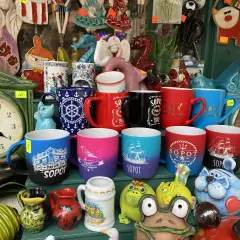 Kolekcja kubków z napisami "Sopot" i morskim motywem, idealne pamiątki z wakacji. W tle zegar i ceramiczne figurki.