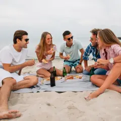Grupa przyjaciół cieszy się piknikiem na plaży, jedząc pizzę i pijąc napoje. To świetny pomysł na wakacje!