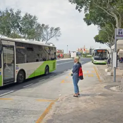 Kobieta czeka na autobus na przystanku przy lotnisku na Malcie. W tle widać inne autobusy i budynki.