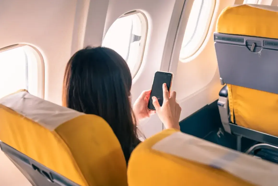 Kobieta w samolocie trzyma telefon, zastanawiając się, **co to tryb samolotowy**. Widok zza jej pleców na żółte siedzenia i okno.