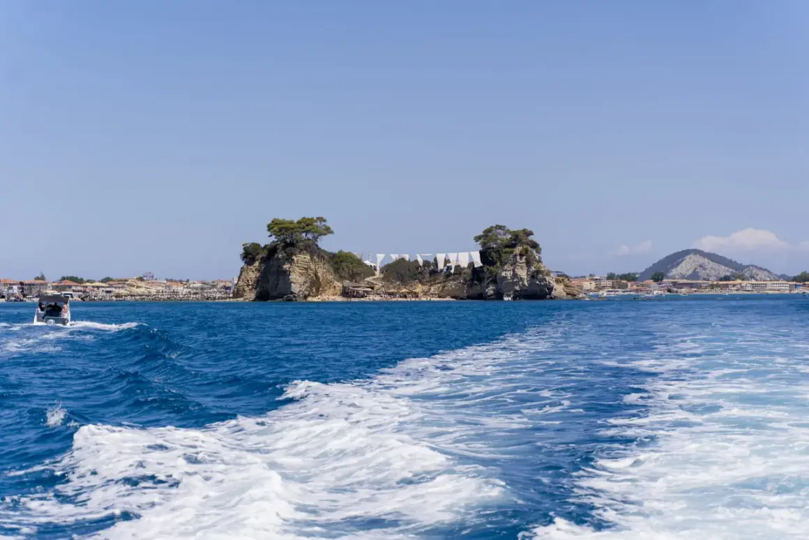 Idealna zakynthos pogoda sierpień: błękitne niebo, turkusowe morze i skaliste wysepki z drzewami. Widać łódź i plażę.