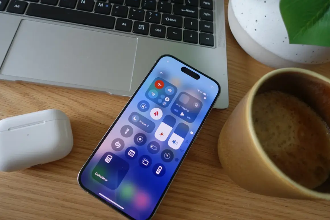 Na ekranie iPhone'a widać centrum sterowania, gdzie łatwo włączyć tryb samolotowy, dotykając ikony samolotu. Obok leży laptop i kubek kawy.