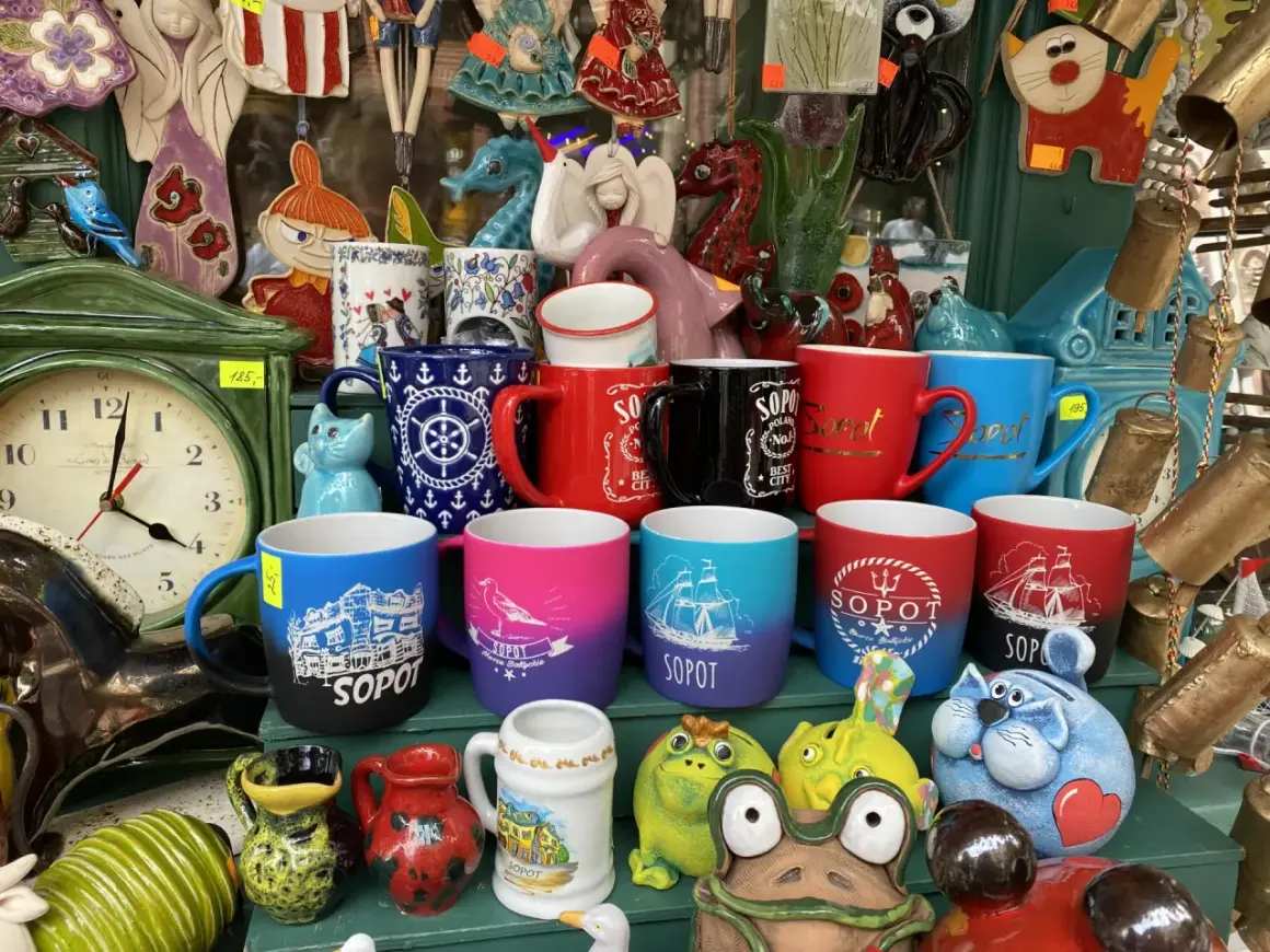 Kolekcja kubków z napisami "Sopot" i morskim motywem, idealne pamiątki z wakacji. W tle zegar i ceramiczne figurki.