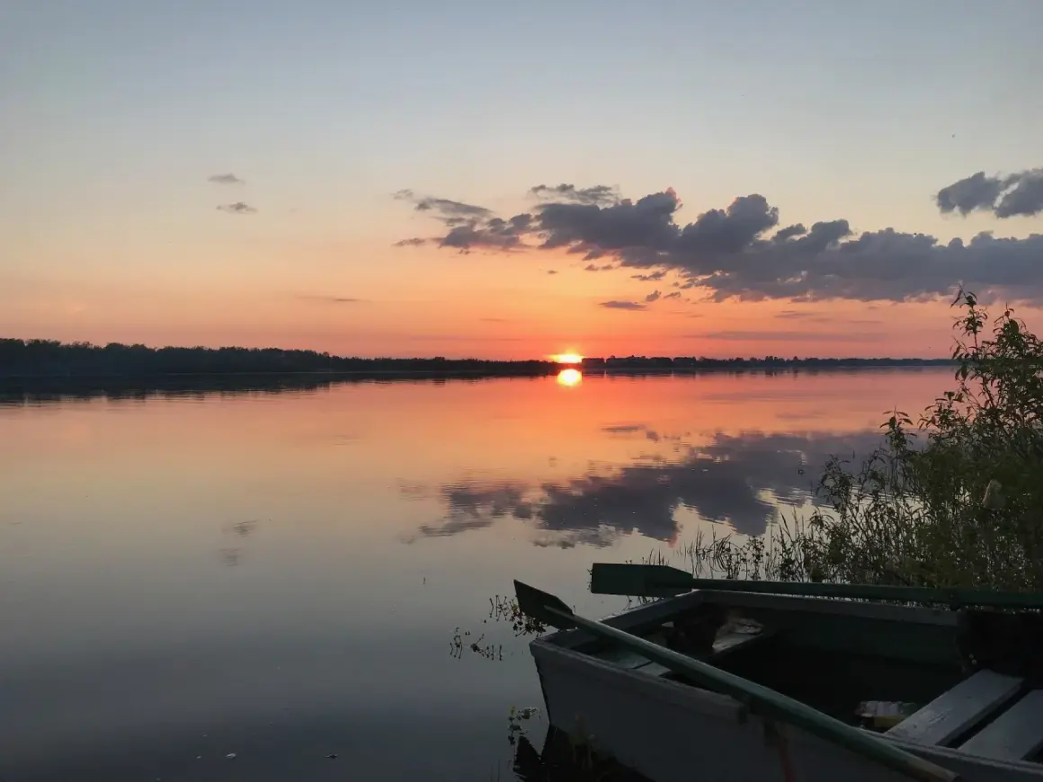 Łódź czeka nad spokojną rzeką białych nocy, odbijając pomarańczowe niebo i chmury.