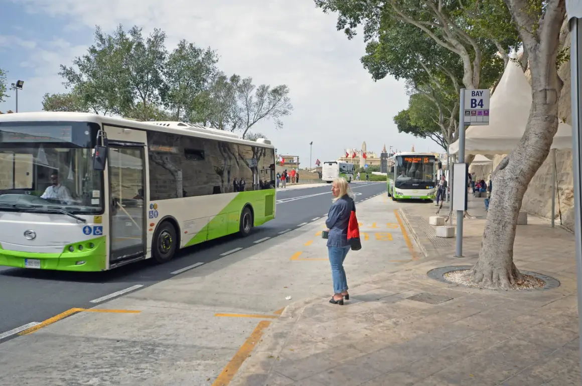 Kobieta czeka na autobus na przystanku przy lotnisku na Malcie. W tle widać inne autobusy i budynki.