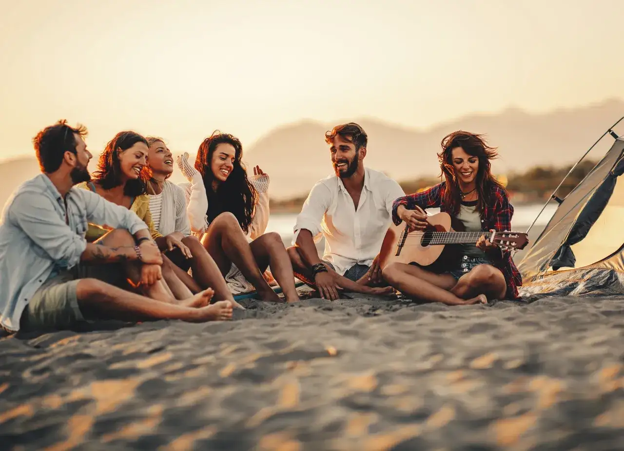 Grupa przyjaciół siedzi na plaży przy namiocie, śmiejąc się i grając na gitarze. Idealne miejsce, by spędzić wakacje w lipcu.
